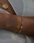 A person wears layered 18k gold plate bracelets, including the Transformed by Love Lotus Bracelet with a lotus flower charm and a pearl accent, plus a detailed gold pendant necklace, all against white textured fabric.