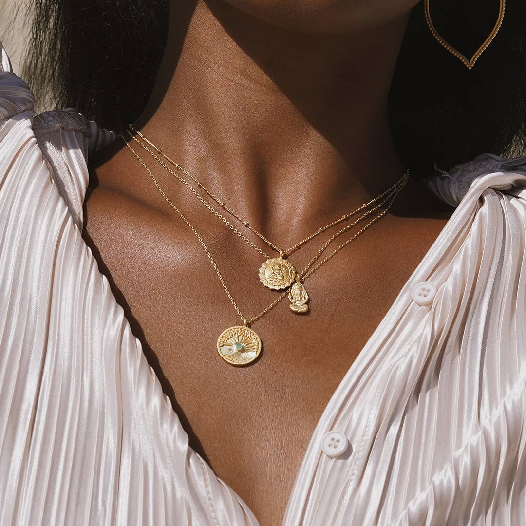 A close-up of a person wearing a pleated, silky white blouse and three layered gold necklaces, including the Abundant Grounding Tree Necklace and other ornate circular charms.