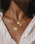 A close-up of a person wearing a pleated, silky white blouse and three layered gold necklaces, including the Abundant Grounding Tree Necklace and other ornate circular charms.