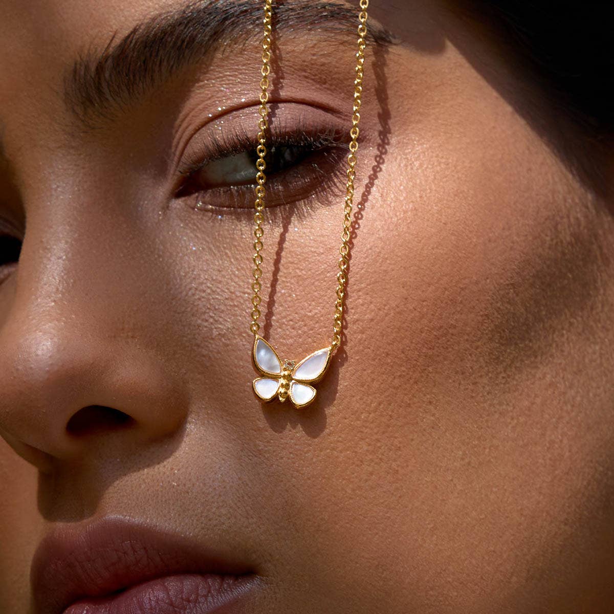 Close-up of a woman's face with smooth skin and natural makeup, holding the Renewed Hope Pearl Butterfly Necklace—delicate 18k gold plated finish—near her eye.