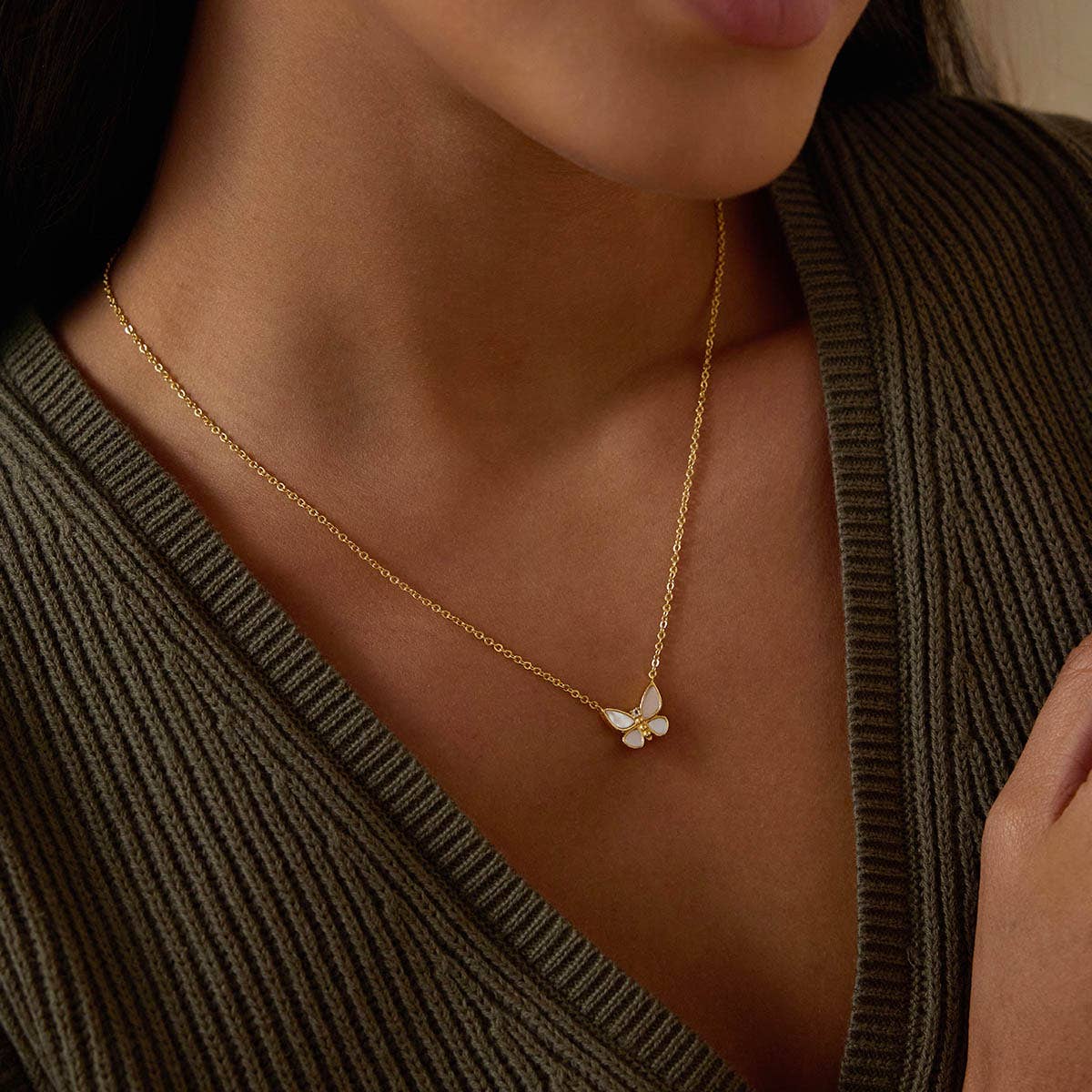 A woman in a ribbed, deep V-neck olive sweater wears the Renewed Hope Pearl Butterfly Necklace—an 18k gold-plated piece featuring a mother of pearl butterfly pendant resting at the base of her neck.