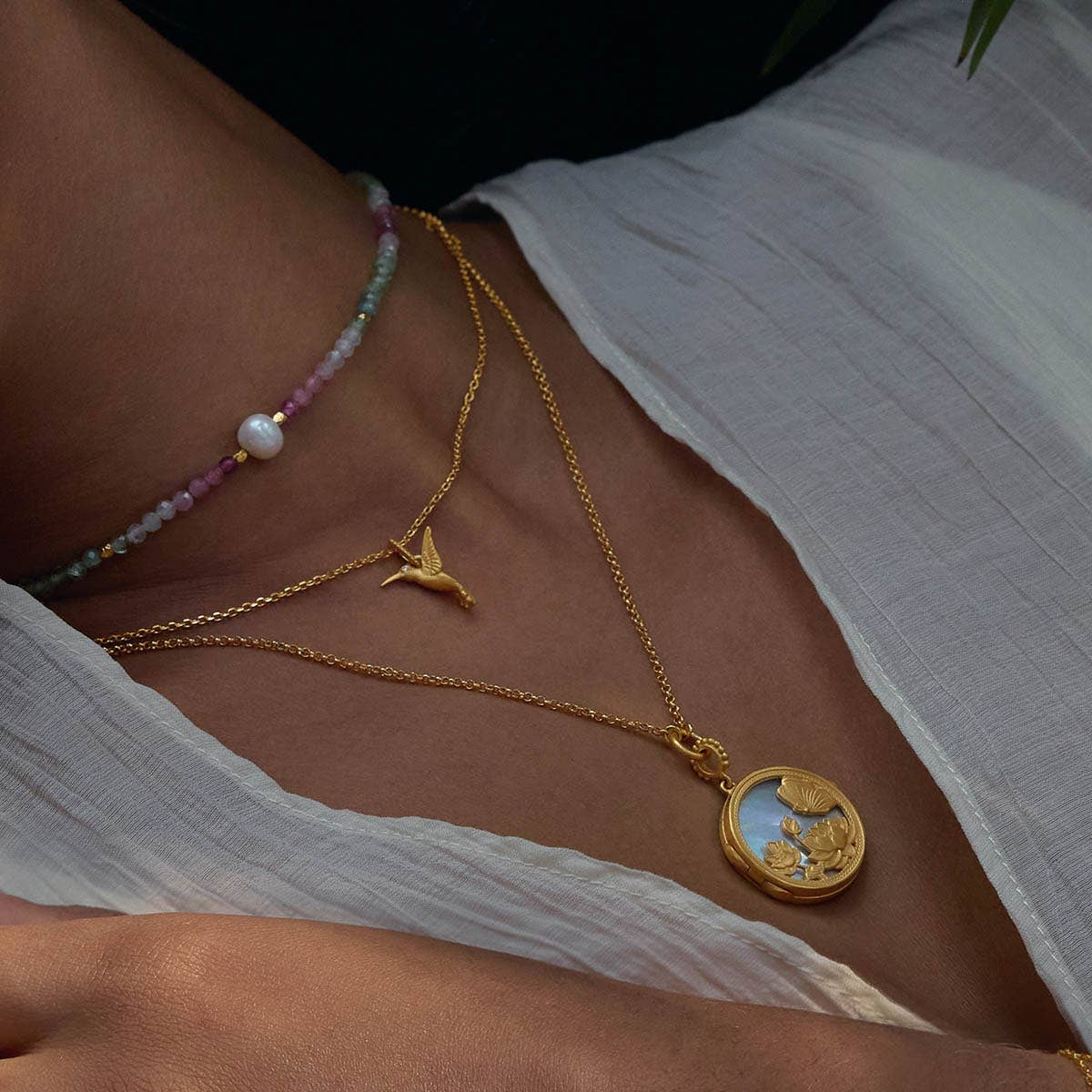 A close-up of a woman in a white textured blouse wears three layered necklaces, including the Loving Devotion Hummingbird Necklace 16&quot;, a multicolored beaded strand, and a large round gold locket with an embossed design.