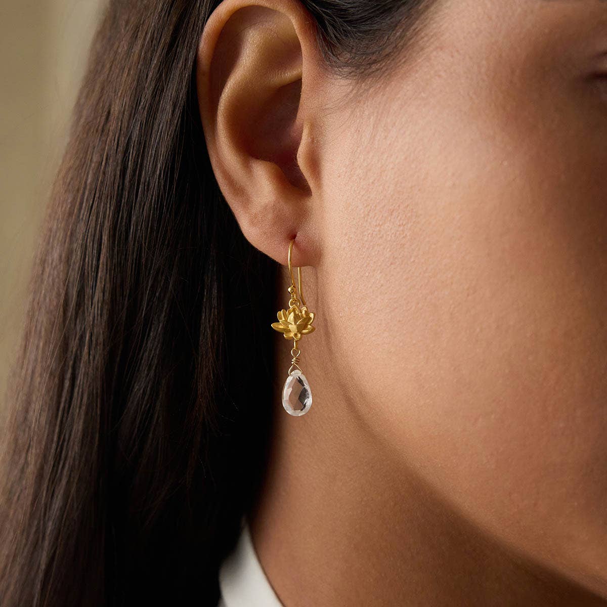 Close-up of a person’s ear wearing the Spiritual Journey Lotus Crystal Earring—an 18k gold plated piece featuring a clear teardrop crystal. Their straight brown hair is tucked back, showcasing this elegant crystal healing jewelry.