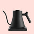 The Matte Black Pro Precision Electric Kettle, with customizable brew settings, a curved spout, and ergonomic handle, sits on its matching square base against a pale pink background.