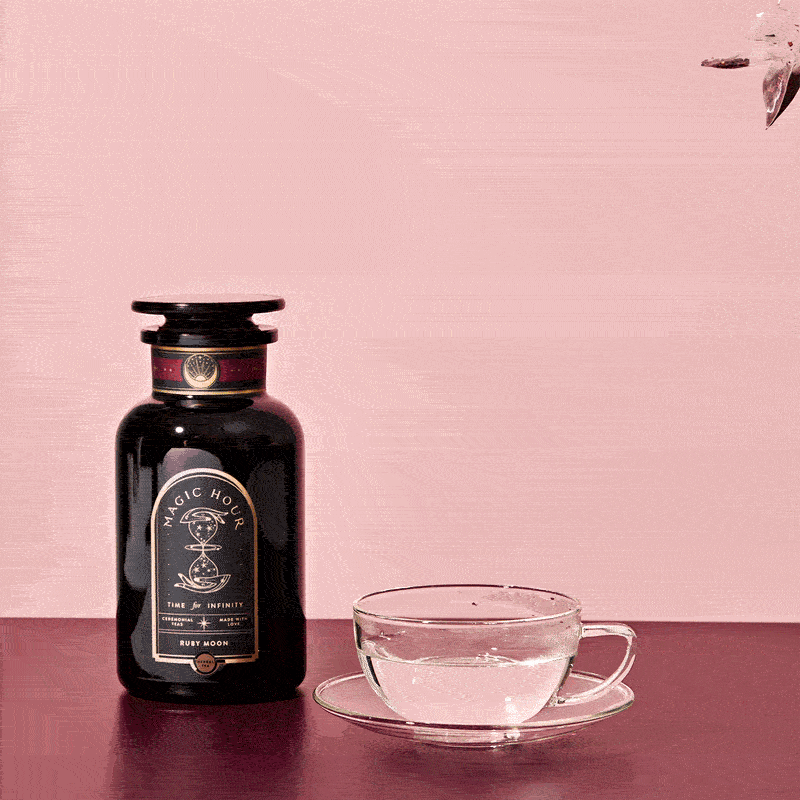 A black bottle labeled "Ruby Moon™ Herbal Tea Sachets" sits on a pink surface beside a transparent glass teacup, as caffeine-free herbal tea is poured into the cup against a pink background.
