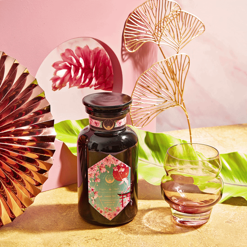 A dark glass bottle labeled "Queen of Kyoto" and a round glass sit on a surface, surrounded by Jasmine Blossoms, pink and gold decor, green leaves, and a reflective fan against a softly vibrant pink and gold background.