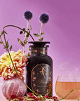 A decorative jar labeled "Libra Shatavari-Cardamom-Rose White Love Cake Tea" sits beside a glass of organic white tea, surrounded by pink and yellow flowers, purple globe thistles, and loose tea leaves on a pinkish-purple background.