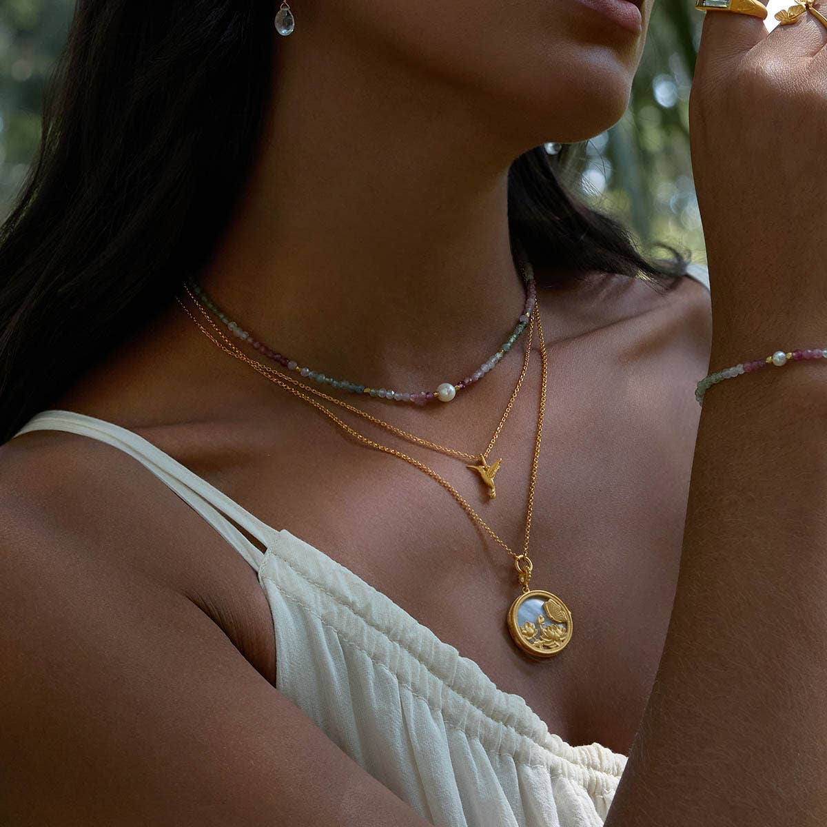 A woman wearing a white off-shoulder top is shown from the shoulders up, accessorized with layered gold necklaces including the Loving Devotion Hummingbird Necklace 16&quot;, plus a beaded bracelet, gold ring, and drop earring in soft natural light.