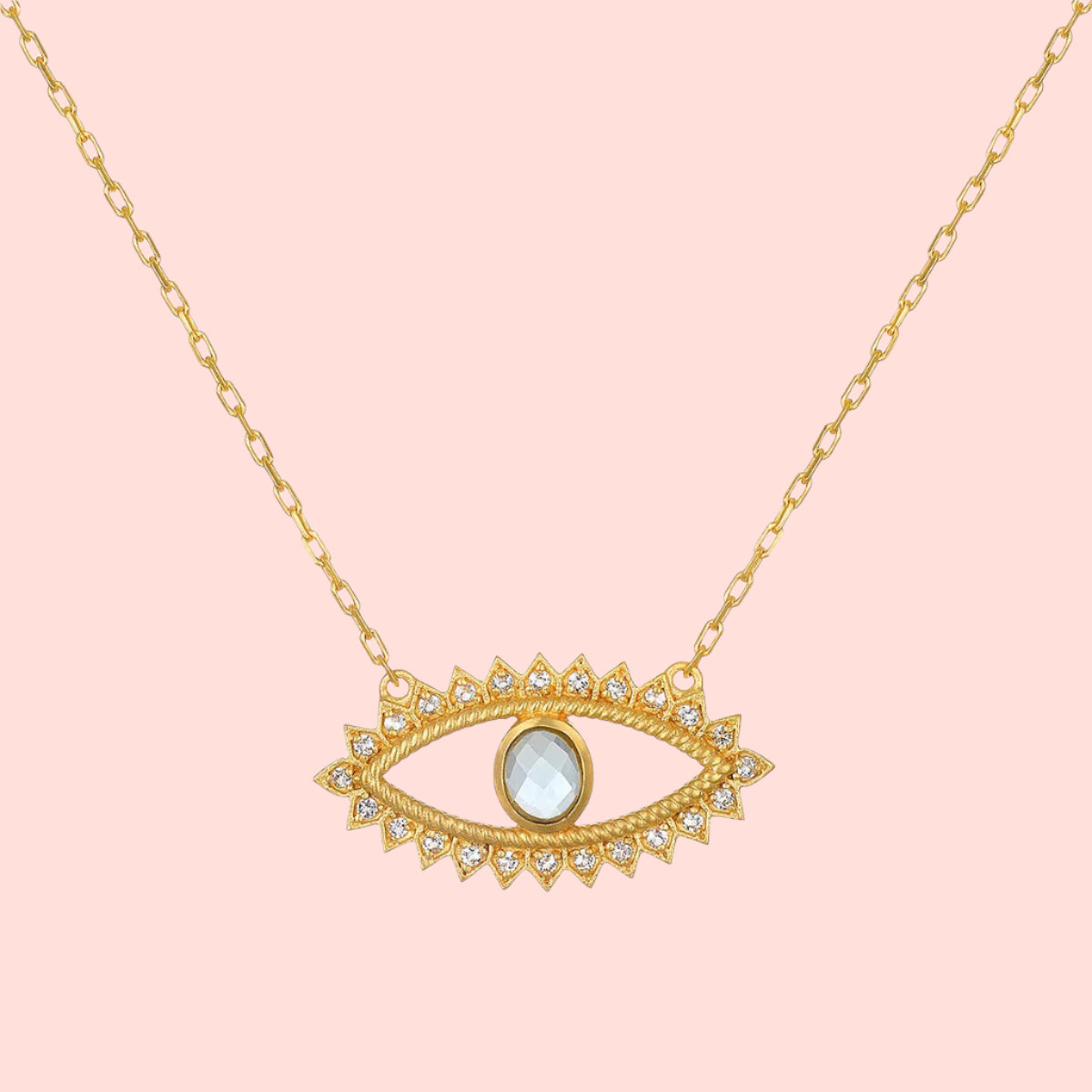 The Eye Blue Topaz Necklace features a gold pendant in an eye shape, accented with clear gemstones and a central blue topaz stone, inspired by the classic evil eye design.