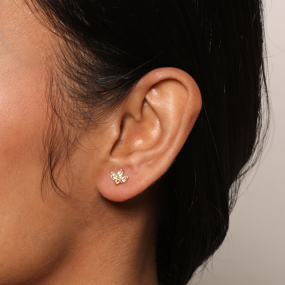 Close-up of a woman's ear wearing White Topaz Lotus Studs—delicate earrings crafted with 18kt gold plate and three sparkling stones—set against a neutral background.