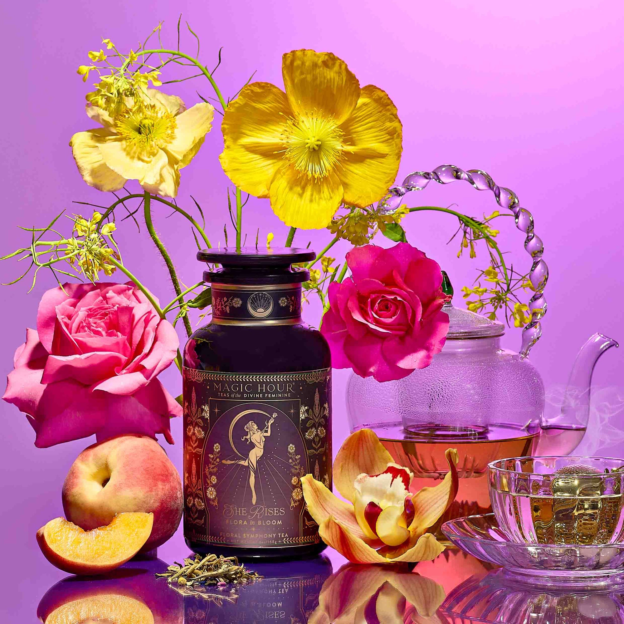 A display featuring the Monthly Magic Tea Journey Subscription—an ornate tea tin, glass teapot, rare botanicals, loose tea leaves, peaches, orchids, roses, and yellow poppies—evokes a goddess ritual on a vibrant pink background.