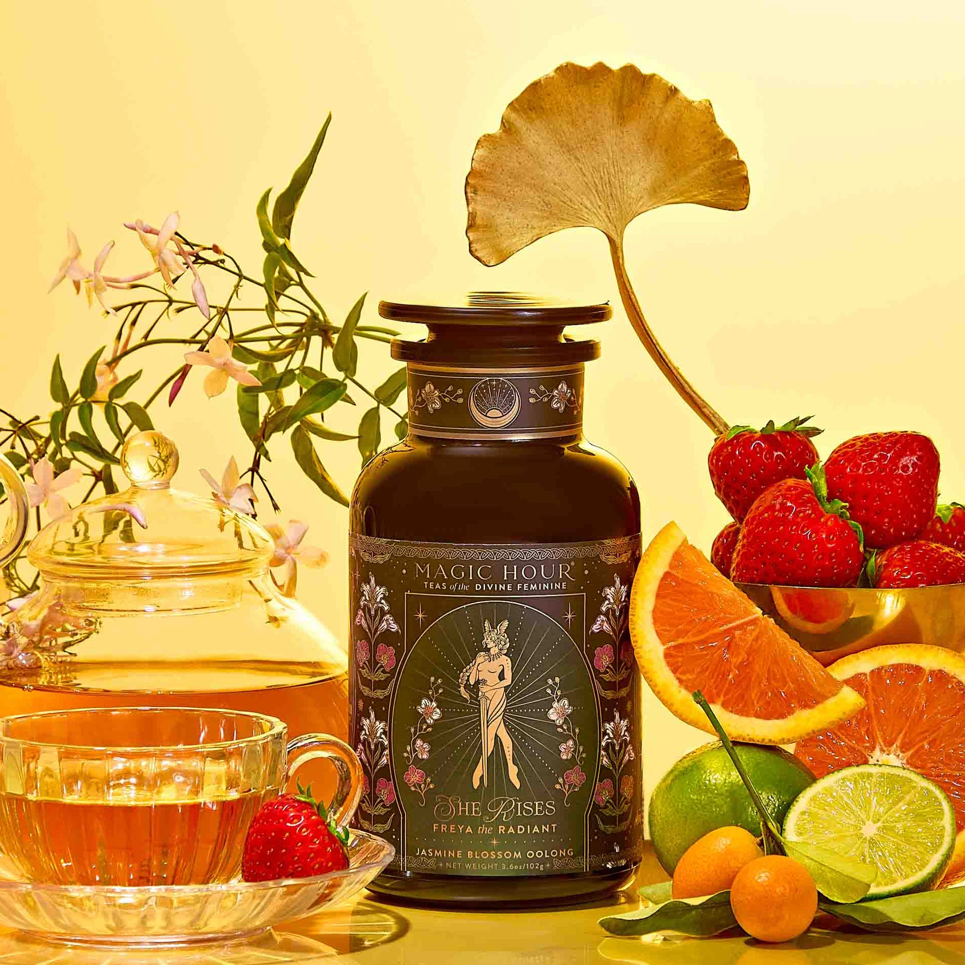 Freya: The Radiant tea set features a glass teapot and wellness tea cup beside a dark floral canister, surrounded by strawberries, orange slices, lime, kumquats, jasmine flowers, and a gold ginkgo leaf on a yellow background.
