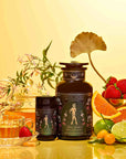A glass teapot and cup filled with Freya: The Radiant oolong sit beside two dark "Magic Hour" jars, fresh strawberries, orange slices, kumquats, and greenery on a vibrant yellow background.