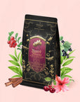 A decorative tea package adorned with gold designs and a red label is surrounded by illustrated berries, green leaves, cinnamon sticks, and a hibiscus flower on a soft pink background—a perfect presentation for Haunted Hibiscus: Tart Berry Herbal Blend with Warm Spices. The imagery evokes the vibrant flavors of this caffeine-free herbal blend, inviting you to indulge in a soothing, aromatic experience.