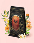 A black and gold tea bag labeled “Candy Corn: Herbal Fall Tea with Vanilla & Caramel”—a delightful caffeine-free blend—is surrounded by yellow flowers, marigolds, and vanilla pods on a light pink background.