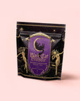 A black and purple bag of “Black Cat Brownie: Decadent Chocolate Puerh Blend with Vanilla & Hazelnut” features gold star designs and illustrations of two celestial women on a pale pink background. This chocolate tea blend offers a magical fusion of flavor and artistry.