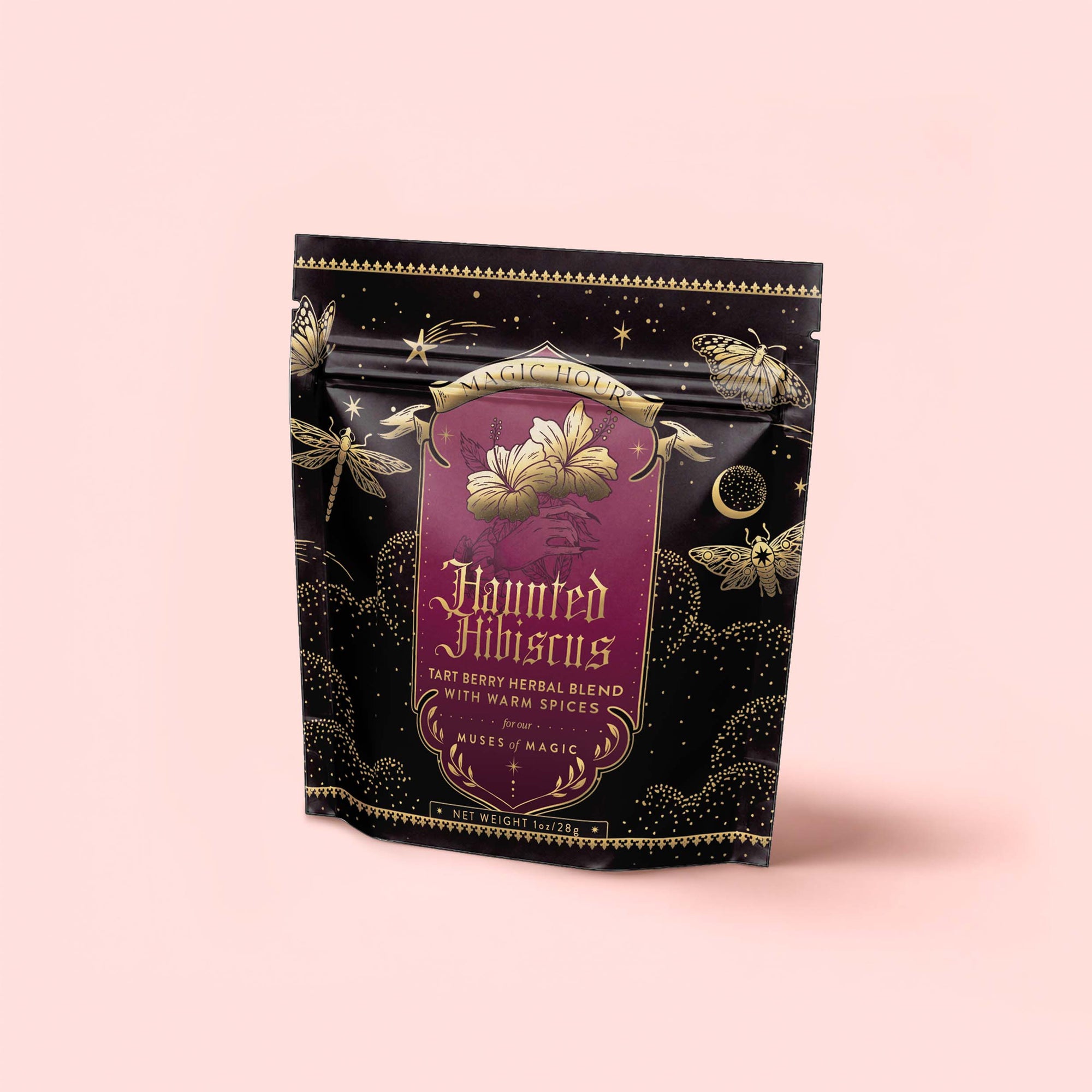A black and gold tea package labeled “Haunted Hibiscus: Tart Berry Herbal Blend with Warm Spices,” adorned with moth and flower illustrations and celestial designs, rests on a pale pink background. This enchanting caffeine-free tea blends tart berries and warm spices for a truly mystical experience.