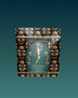 A decorative black tea bag features gold floral patterns and an illustration of a woman with radiant rays. The label reads “Magic Hour Hathor of Joy: Honey Nectar Herbal Tea.” The background displays a gradient of teal.