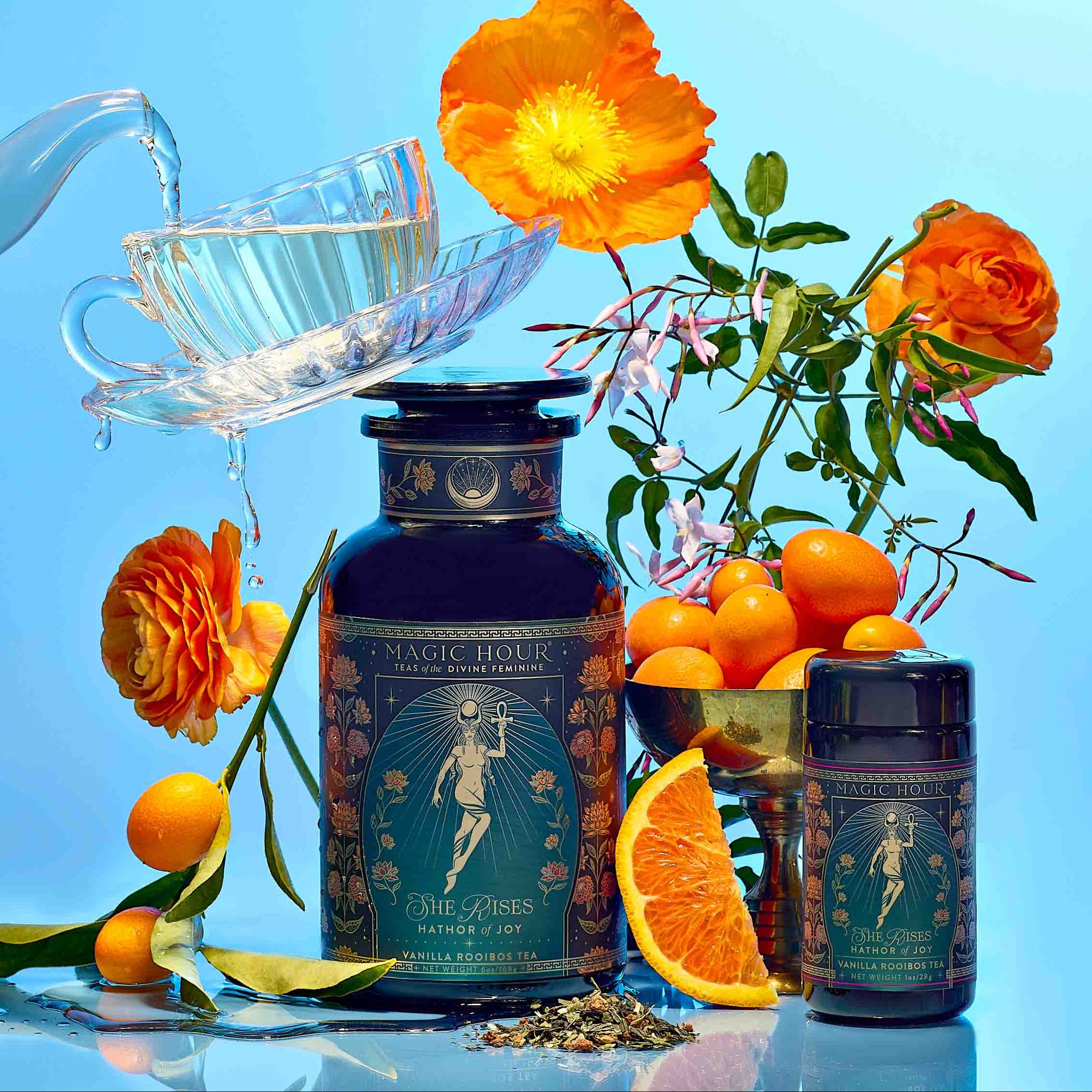 A teacup is being filled with tea beside two "Monthly Magic Tea Journey Subscription" containers—ideal for a goddess ritual—surrounded by orange flowers, kumquats, an orange slice, and loose tea leaves against a vibrant blue background.
