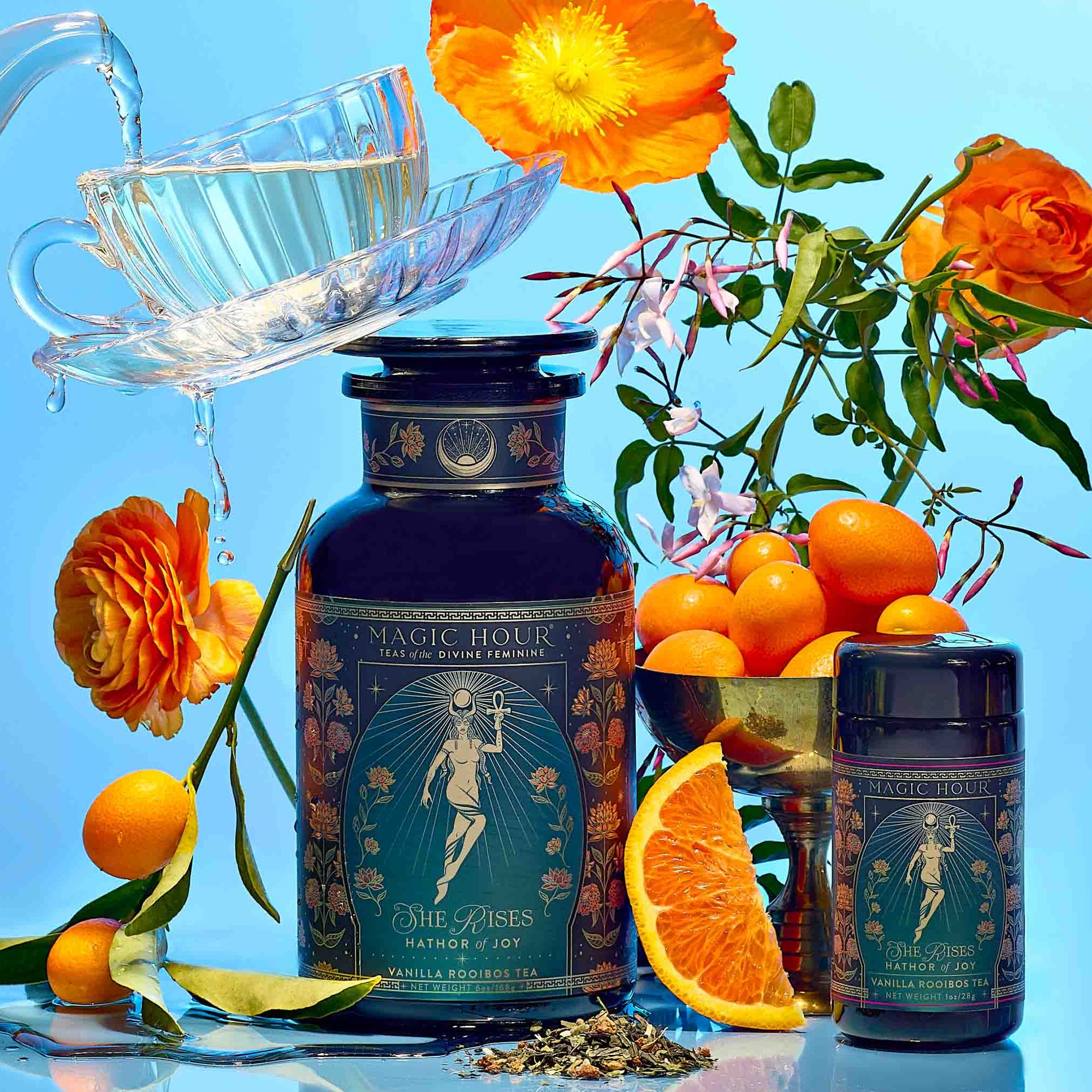 Hathor of Joy: Honey Nectar Herbal Tea is poured into a teacup beside a large ornate tea jar, small tin, kumquats, orange slice, tea leaves, and orange flowers on a vibrant blue background.