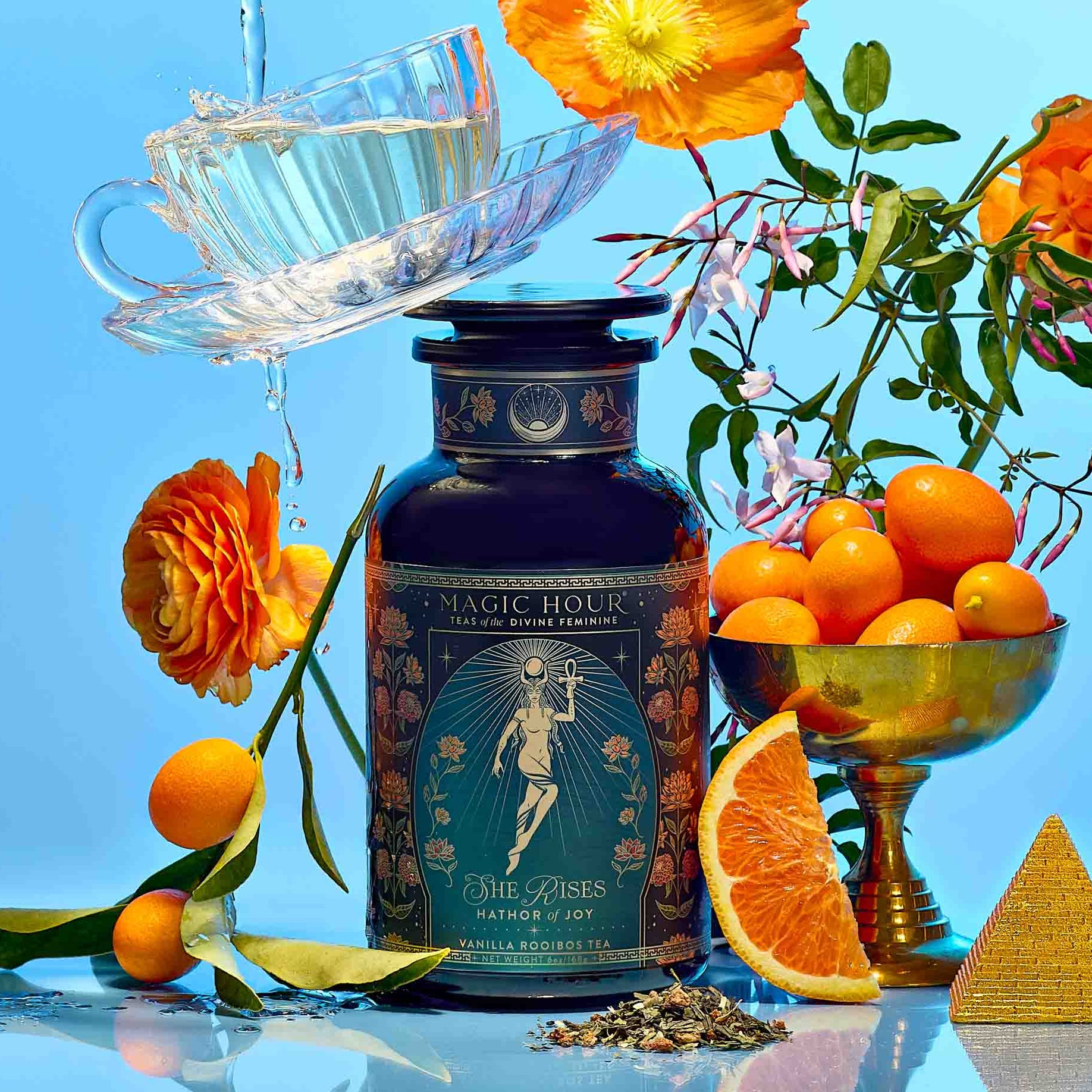 A bottle of Hathor of Joy: Honey Nectar Herbal Tea is surrounded by sliced citrus fruits, orange kumquats in a gold bowl, orange flowers, and a teacup with water splashing out against a vibrant blue background.
