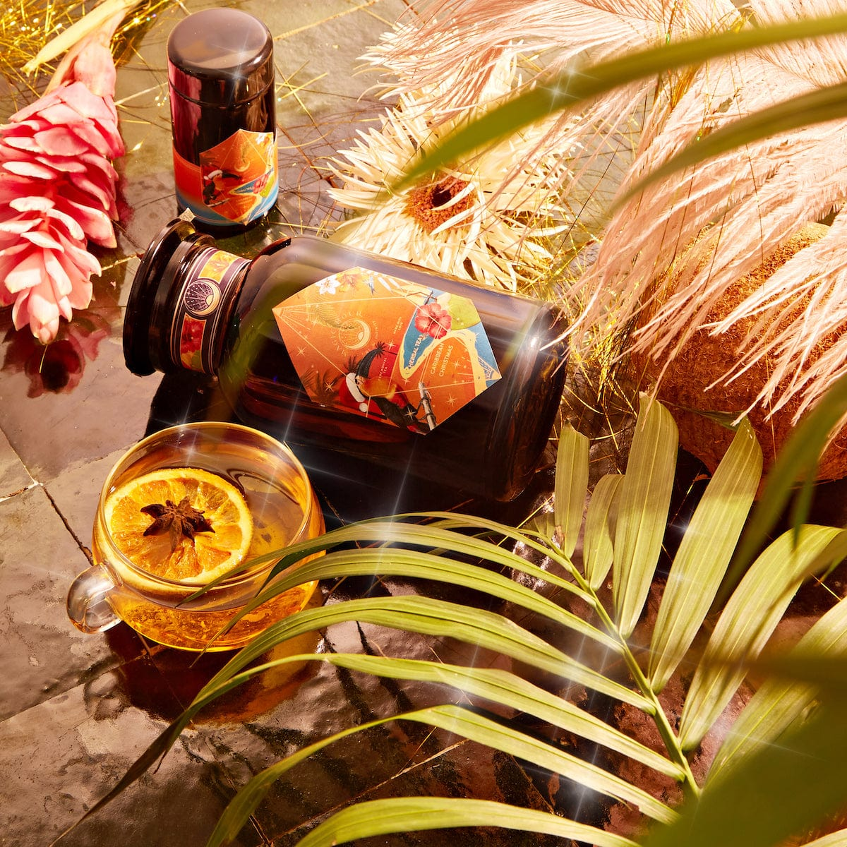 A glass cup of tea garnished with a slice of orange and a star anise is placed on a reflective surface surrounded by tropical leaves. Nearby, there's a large bottle and a smaller container, both with vibrant labels. A pink tropical flower is also visible.