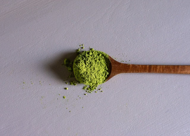 A wooden spoon lies on a flat surface, filled with bright green matcha powder. Some of the powder has spilled onto the surface around the spoon. The background is a neutral light gray.