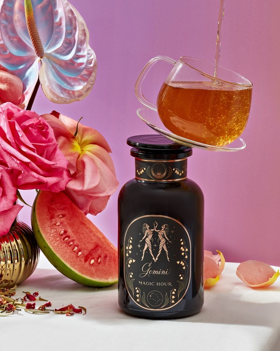 Tea being poured into a glass cup balanced on a Gemini Astrology Tea Gift Set bottle, set among flowers and a slice of watermelon.