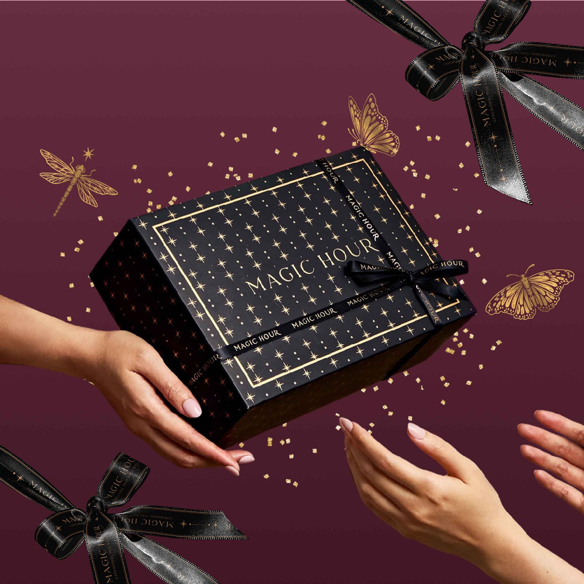 A black gift box featuring gold stars and “Magic Hour” text, decorated with black ribbon bows, gold confetti, butterflies, and dragonflies on a maroon background is passed between two sets of hands—making it the perfect way to present the Labor Day Mystery Box, a seasonal collection of exclusive teas.