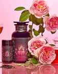 The Monthly Magic Tea Journey Subscription is artfully displayed alongside a glass of tea, lush pink roses, and a teapot, all set against a dreamy pink backdrop. The packaging showcases an elegant design adorned with a woman and gold accents—ideal for those looking to elevate their daily goddess ritual or connect with the essence of the Divine Feminine.