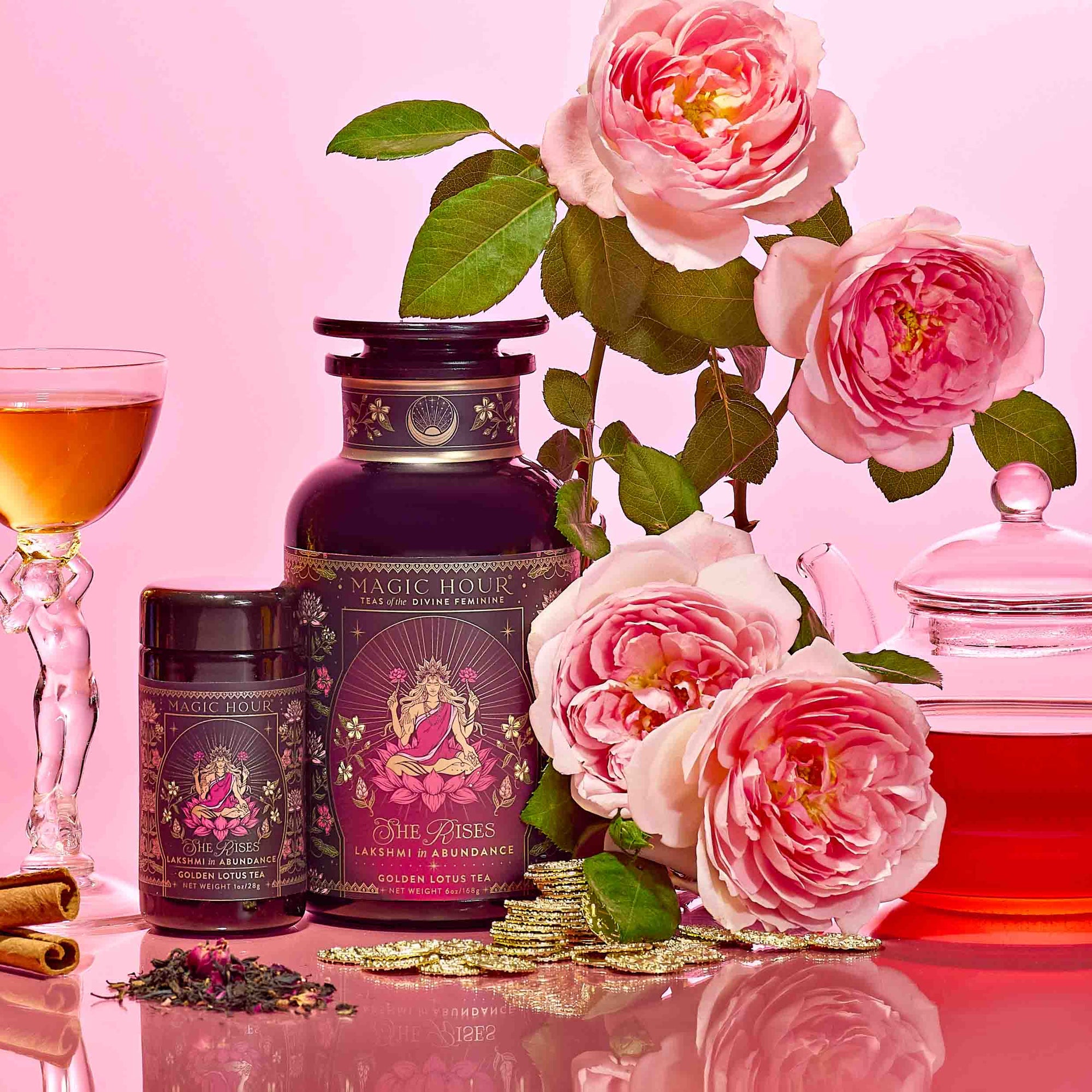 Bottles of Lakshmi in Abundance: Rose Saffron Black Tea for Good Fortune & Radiance with pink labels, a glass of this luxurious blend, a teapot, and blooming pink roses arranged on a pink surface with gold beads evoke the essence of radiant black tea and the delicate charm of saffron rose.