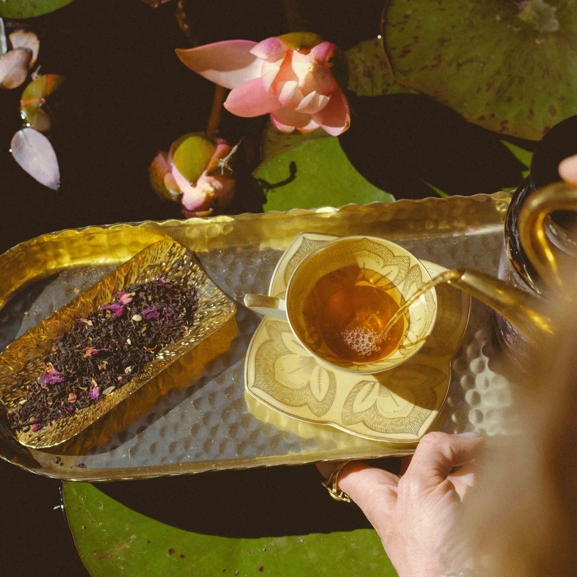 A hand pours tea from the Monthly Magic Tea Journey Subscription into a patterned teacup on a gold tray beside loose tea leaves, with pink lotus flowers and green lily pads floating in water in the background—an invitation to a goddess ritual that honors the Divine Feminine with every soothing sip.