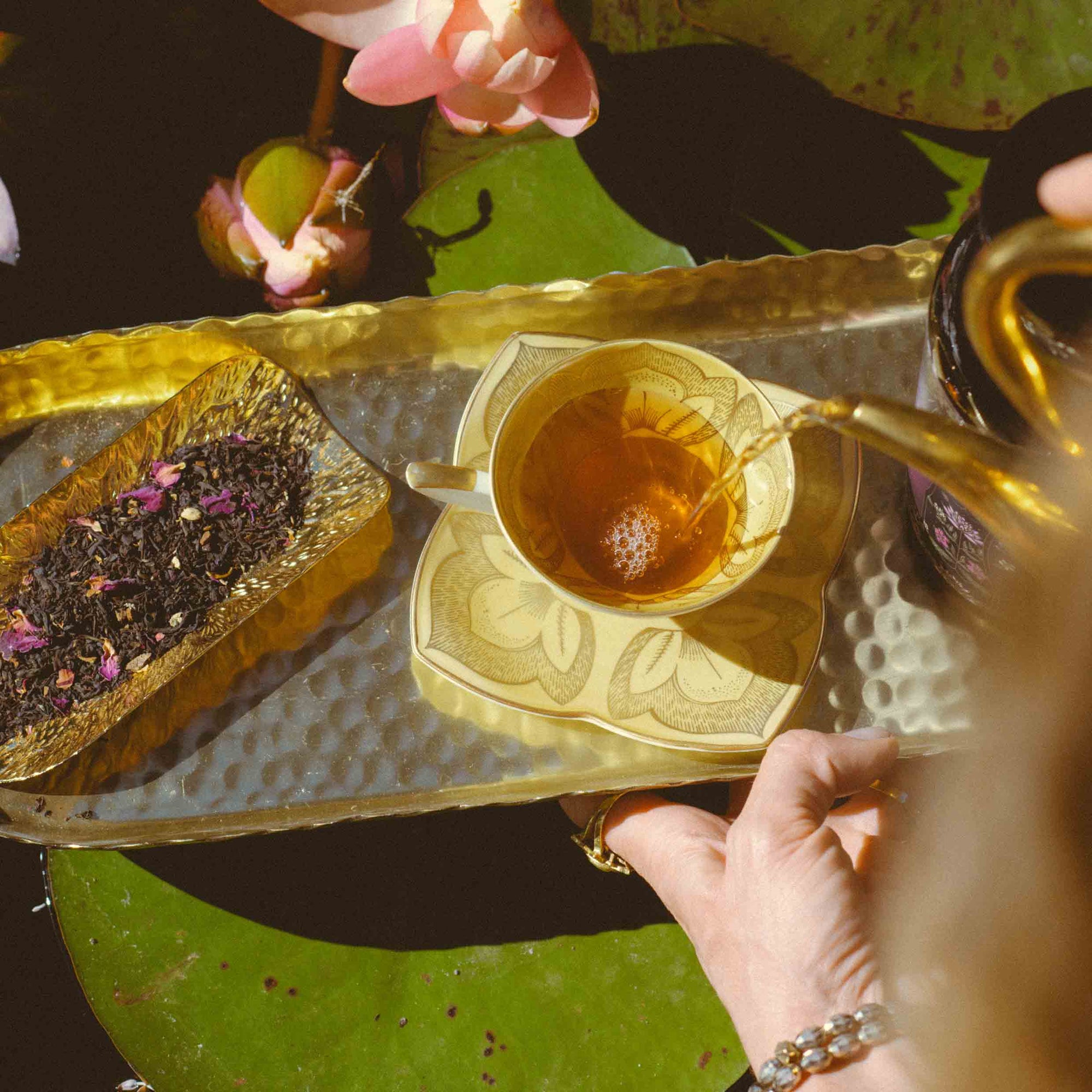 A hand pours Lakshmi in Abundance: Rose Saffron Black Tea for Good Fortune &amp; Radiance into a yellow floral teacup set on a gold tray with loose tea leaves and petals, surrounded by blooming lotus flowers and green lily pads.