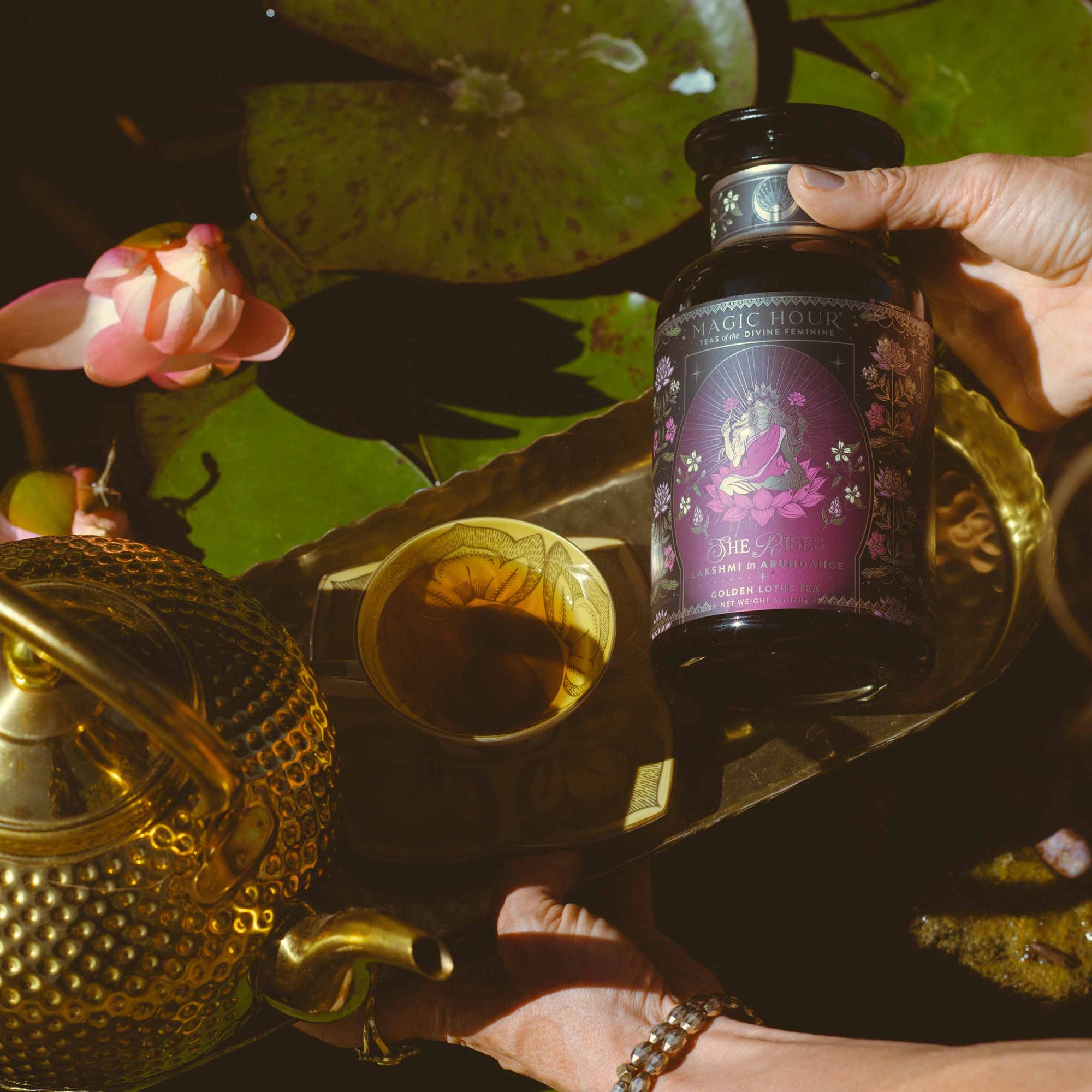 A hand holds a jar of Lakshmi in Abundance: Rose Saffron Black Tea for Good Fortune &amp; Radiance above a brass tray with a teapot and cup. Pink lotus flowers and lily pads grace the background, setting the scene for savoring this antioxidant-rich, luxury black tea.