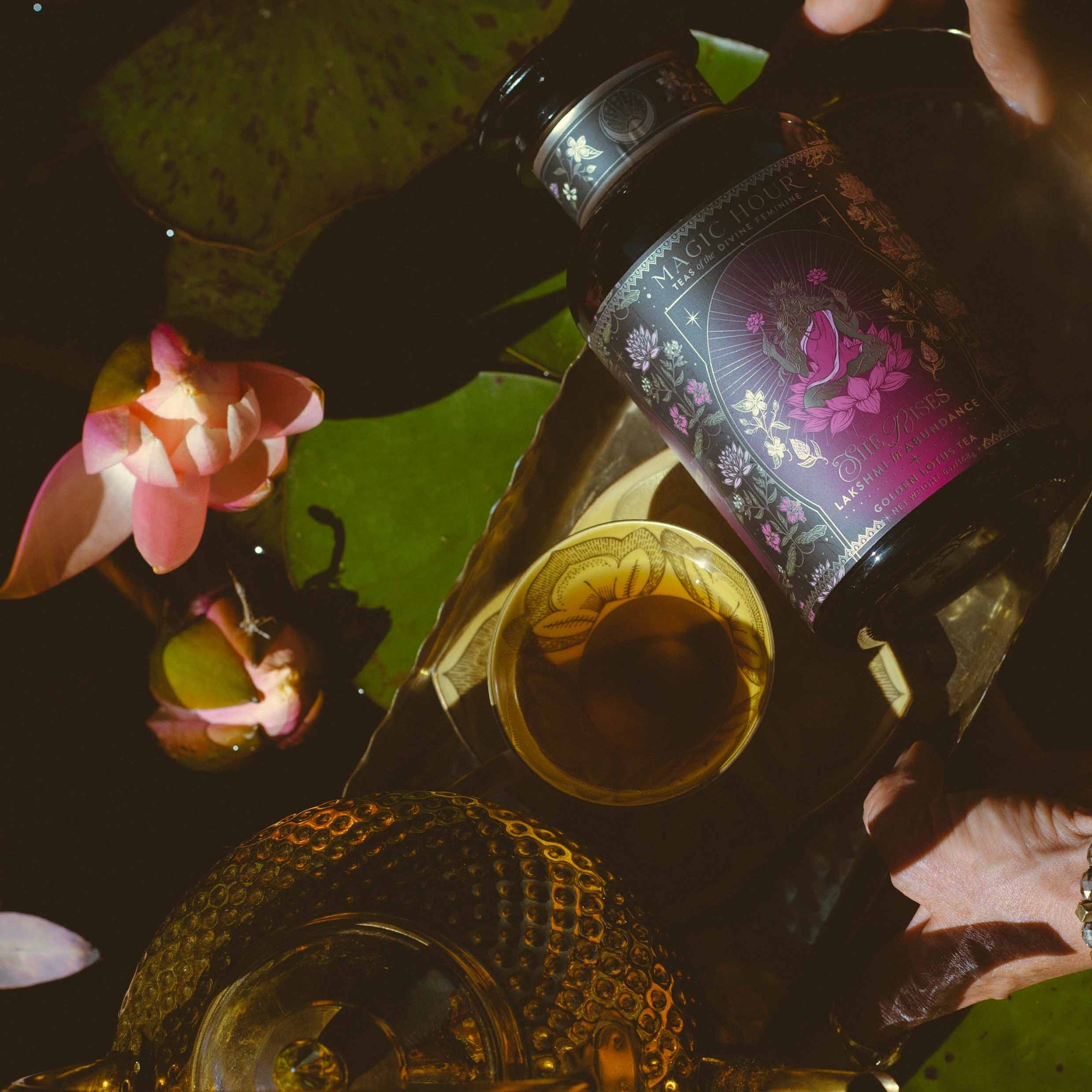 A hand holds a jar of Monthly Magic Tea Journey Subscription tea with a purple floral label next to a teacup, teapot, water lily flowers, and lily pads, all bathed in warm sunlight—perfect for a goddess ritual celebrating the Divine Feminine.