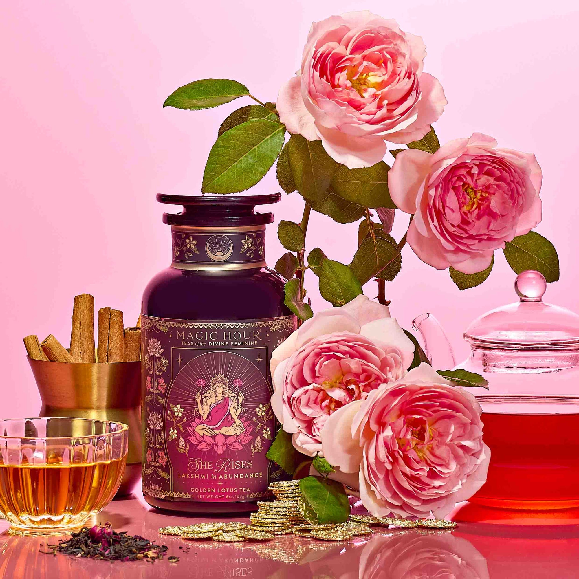 A tea tin labeled “She Rises” sits beside a glass teacup, teapot, cinnamon sticks, loose luxury black tea, and blooming pink roses on a pink background—inviting you to indulge in the radiance-boosting Lakshmi in Abundance: Rose Saffron Black Tea for Good Fortune &amp; Radiance experience.