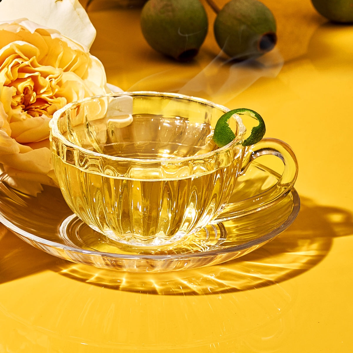 A London Fog Tea Sampler Set brew in a glass teacup with a green garnish sits on a saucer atop a yellow surface beside a yellow rose. Blurred green fruits are in the background as steam softly rises from the cup.