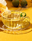 A London Fog Tea Sampler Set brew in a glass teacup with a green garnish sits on a saucer atop a yellow surface beside a yellow rose. Blurred green fruits are in the background as steam softly rises from the cup.