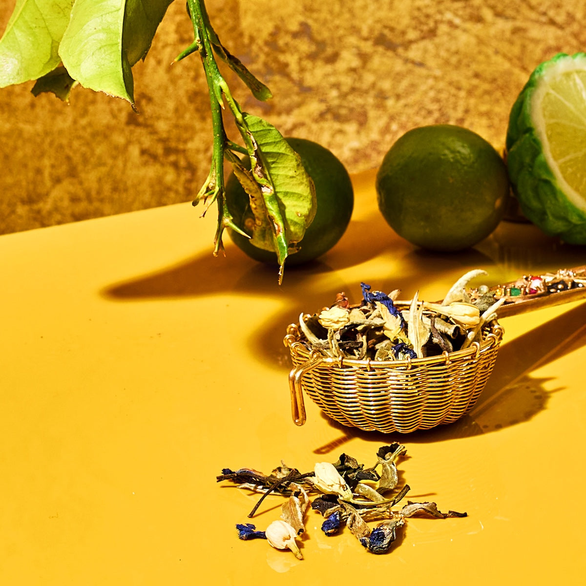 A gold tea infuser filled with dried herbs and blue petals rests on a yellow surface, some herbs spilled out. In the background, a branch with green leaves and two whole limes, one partially sliced, evoke the flavors of Earl Grey in the London Fog Tea Sampler Set.