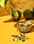 A gold tea infuser filled with dried herbs and blue petals rests on a yellow surface, some herbs spilled out. In the background, a branch with green leaves and two whole limes, one partially sliced, evoke the flavors of Earl Grey in the London Fog Tea Sampler Set.