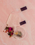 Two mesh tea bags lie arranged on a pink marble surface. One bag is partially filled with a vibrant blend of loose tea leaves and dried petals spilling out, while the other remains empty. Tags attached to the bags read "Magic Hour." These artisan-crafted strainers from the Magic Hour Starter Kit elevate any tea ceremony with grace and ease.