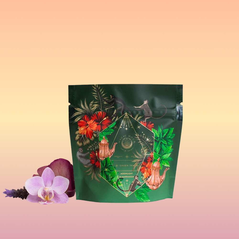 A stand-up pouch in a vibrant shade of green, adorned with intricate floral and animal designs, including flowers and a geometric pattern, is set against a clean white backdrop. Beside the pouch lies a delicate arrangement of pink and purple flowers, capturing the essence of Moroccan mint tea with subtle hints of organic ingredients. The pouch features "Moonlight in Marrakesh: Spiced Mint Green Tea Case Packs" from the brand Magic Hour.