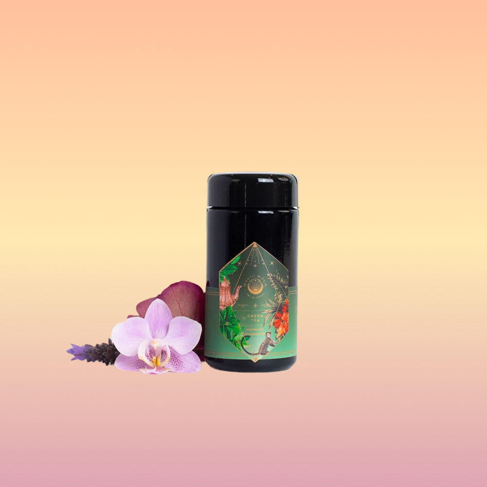 A Moonlight in Marrakesh: Spiced Mint Green Tea case pack by Magic Hour, with its black cylindrical container and decorative green and gold design, stands on a white surface. Surrounding it are pink and purple orchids, lavender flowers, and hints of Moroccan mint tea, adding a touch of nature and color to the scene.