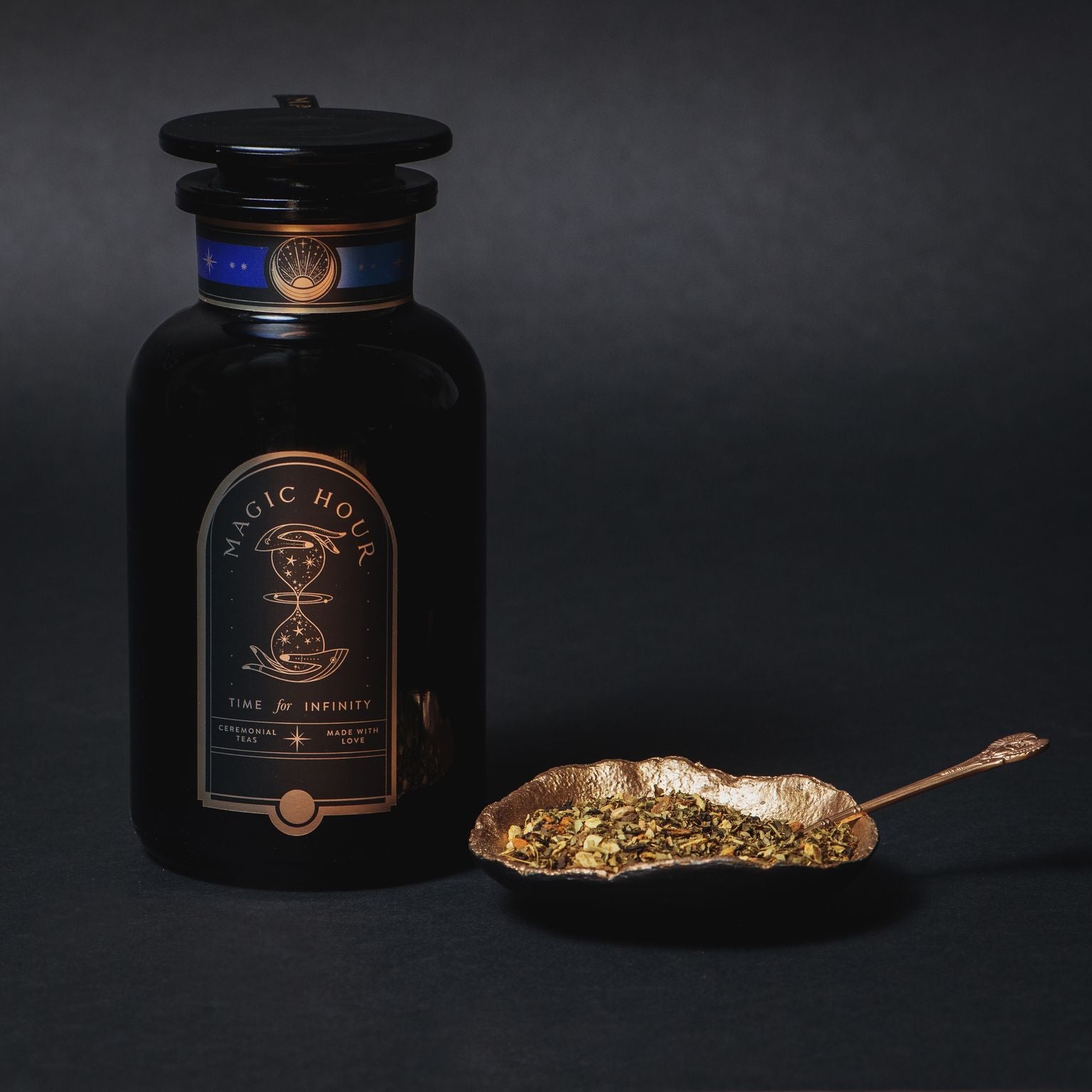 A black jar labeled "Moringa Moon Herbal Tea" sits beside a gold leaf-shaped spoon filled with loose detox tea leaves, all set against a dark background.