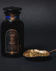 A black jar labeled "Moringa Moon Herbal Tea" sits beside a gold leaf-shaped spoon filled with loose detox tea leaves, all set against a dark background.