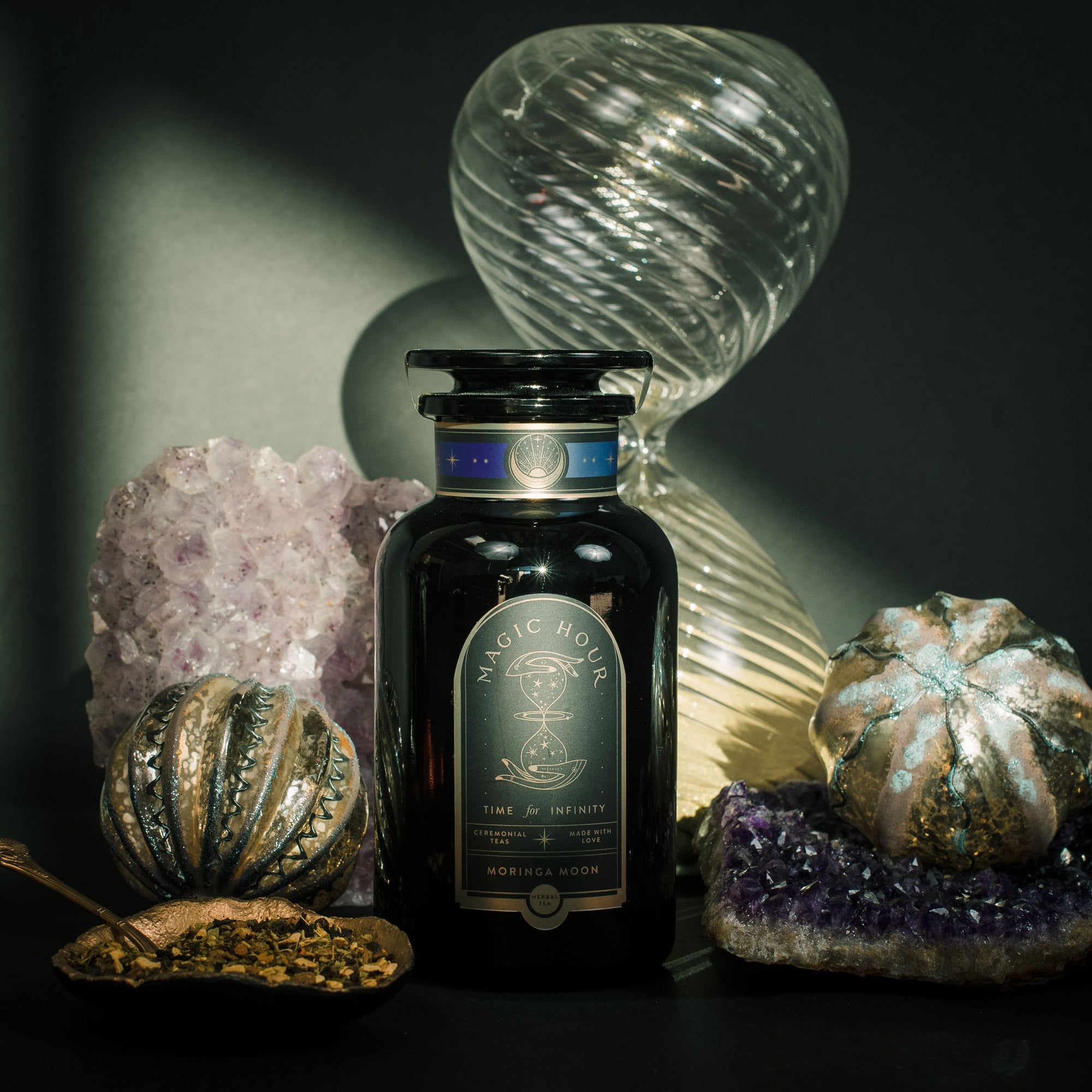 A dark glass jar labeled "Moringa Moon Herbal Tea" stands on a table with amethyst crystals, gourd-shaped glass ornaments, and a spoonful of loose caffeine-free tea, illuminated by dramatic lighting and shadows.