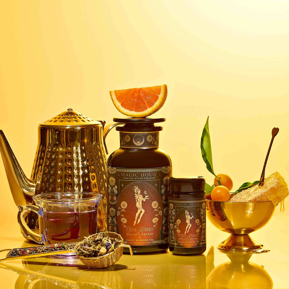 Oshun’s Nectar: Honeyed Herbal Tea for Joy &amp; Radiance is artfully arranged with a teapot, tea cup, glass jar labeled with a fairy and topped with half a grapefruit, a smaller jar, fresh kumquats with leaves, honeycomb, and loose golden honeyed herbal tea on a vibrant yellow background.