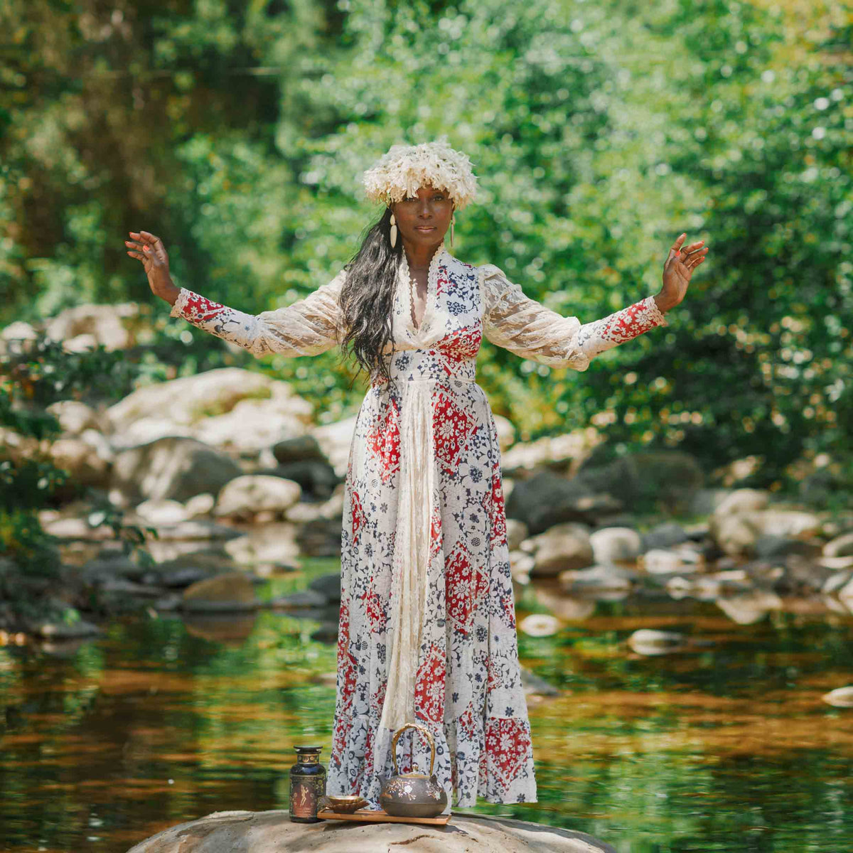 A person in a floral dress and flower crown stands with arms raised on a rock by a stream, surrounded by greenery. At their feet rests a teapot of Oshun’s Nectar: Honeyed Herbal Tea for Joy &amp; Radiance and a jar.