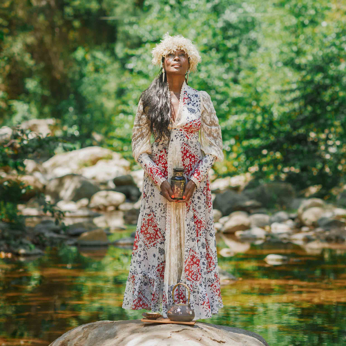 A woman in a long floral dress and flower crown stands on a rock by a stream, holding her Monthly Magic Tea Journey Subscription, surrounded by greenery and sunlight—an embodiment of goddess archetypes, guiding an enchanting ritual in nature.