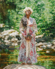 A woman in a long floral dress and flower crown stands on a rock by a stream, holding her Monthly Magic Tea Journey Subscription, surrounded by greenery and sunlight—an embodiment of goddess archetypes, guiding an enchanting ritual in nature.